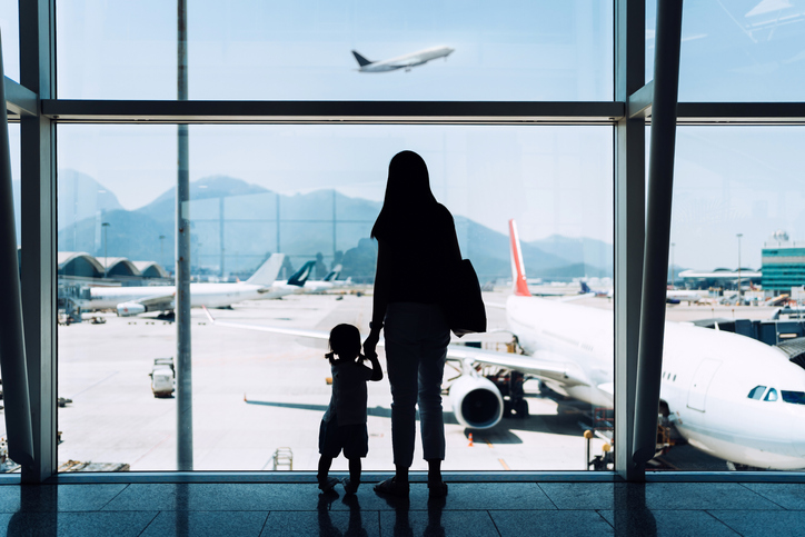 Silhouette of joyful young Asian mother holding hands with cute little daughter looking at airplane through window at the airport while waiting for departure