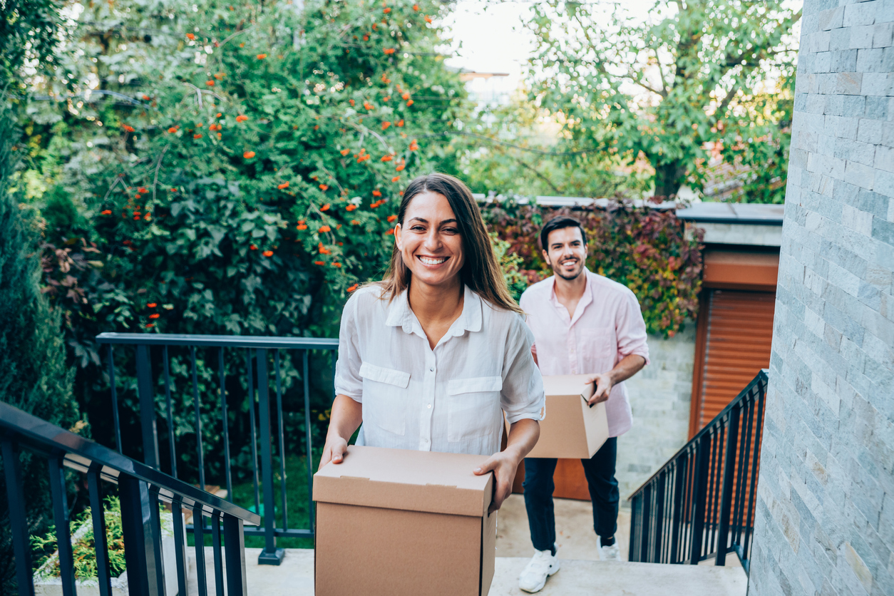 Portrait of happy young couple moving to new house with cardboard boxes. Couple move into their new home. Smiling couple carrying boxes into new home on moving day. Young family entering in their new house.