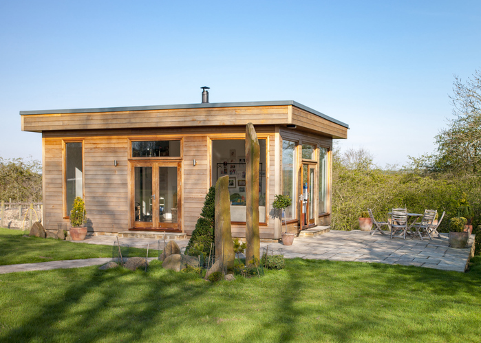 A wide view shot of a modern garden office on a bright summers day, a clear sky is above the wooden office and standing stones can be seen on the grass.