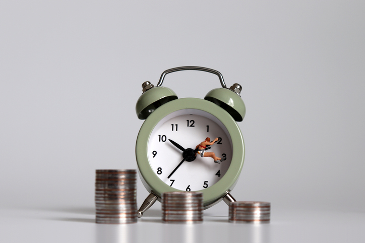 A miniature person with an alarm clock and a pile of coins. A concept of working hours and income inequality.