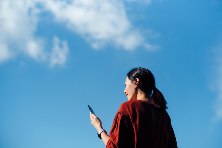 Low angle portrait of young Asian woman using smartphone against beautiful blue sky with cloudscapes, enjoying sunlight outdoors. Lifestyle and technology