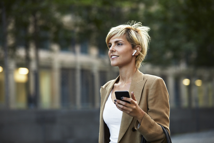 Confident beautiful young businesswoman holding smart phone while looking away. Blond female entrepreneur is walking in downtown district. She is wearing blazer in city.